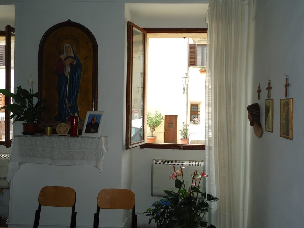 A bright room with a small altar holding a statue of Our Lady, an open window, two chairs, and potted flowers.