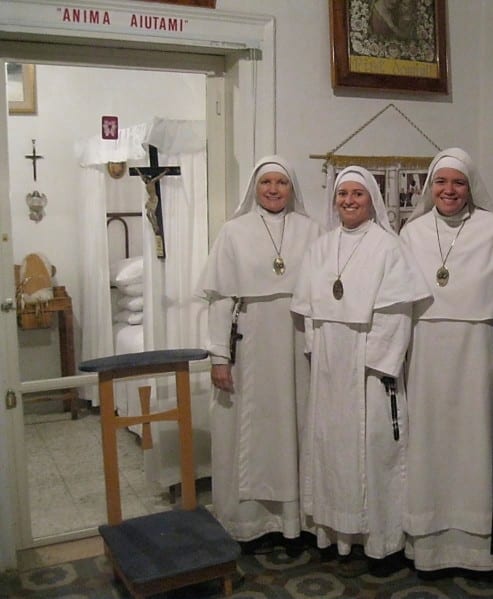Three sisters in white habits smiling beside a wooden chair inside a small room; a sign reading "Anima Aiutami" is visible on the wall above.