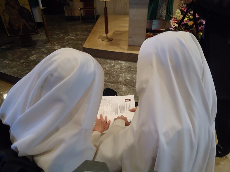 Two sisters in white veils kneeling side by side in a church, reading from a text together.
