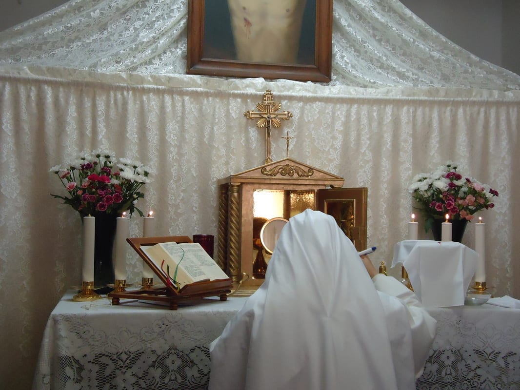 A sister in a white habit kneeling in deep prostration before a small altar with an open tabernacle, candles, flowers, and an open lectionary, with a Sacred Heart image on the wall above.