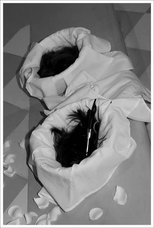 Black-and-white close-up of two white cloth slippers resting among scattered rose petals.