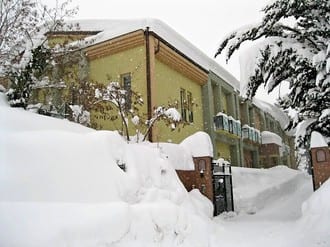 A yellow building under deep snow cover in winter, with heavily snow-laden trees and shrubs filling the foreground.