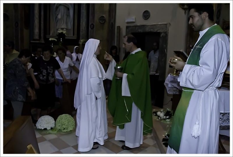 A sister in a white habit receiving a blessing or greeting from a priest in green vestments inside a church.