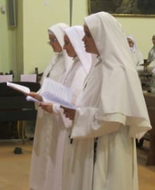 Three sisters in white habits standing in a church or chapel, holding an open booklet and singing or reading aloud.