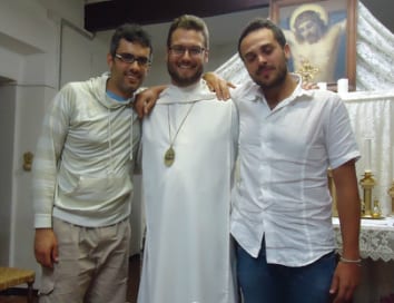 A priest in white vestments with two young men, one on each side, standing in front of a shrine or chapel altar and smiling.