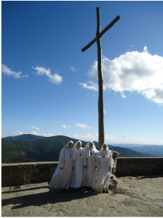 A group of sisters in white habits gathered tightly around the base of a large wooden cross on a hilltop or terrace, with a broad mountain landscape behind them.