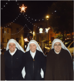 Three sisters in dark winter coats and white veils standing together outdoors at night beneath festive Christmas lights strung across a town square.