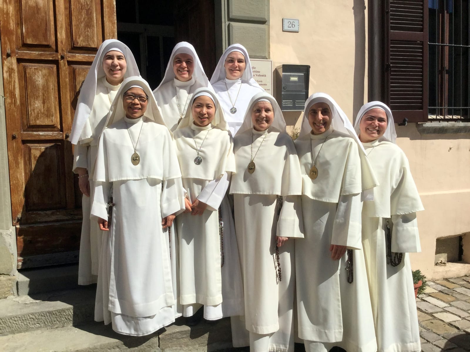 Eight sisters in white habits and veils standing together in a sunlit stone doorway.
