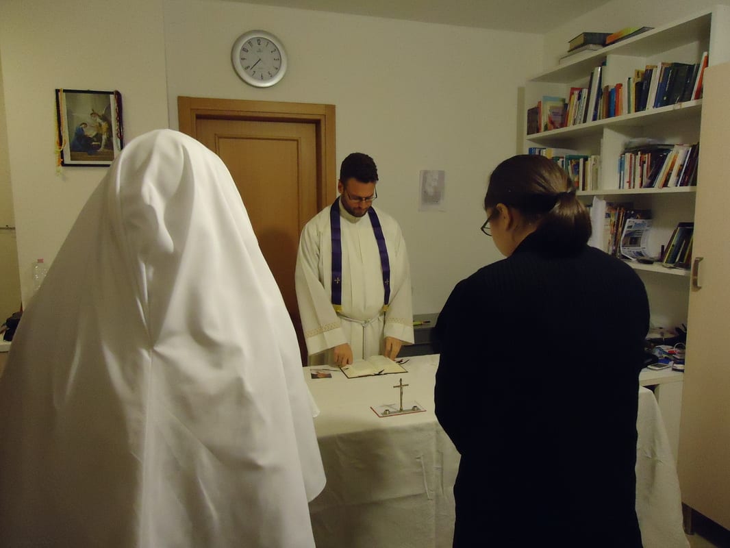 A sister in a white habit and a woman in civilian clothing standing before a priest at a table laid as an altar in a small room.