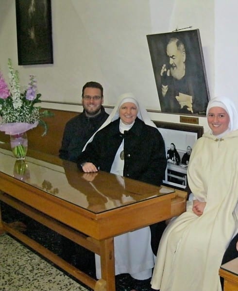 Two sisters and a man seated at a glass-topped table in a room with a large framed photograph of Padre Pio on the wall behind them.