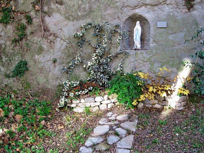 An outdoor stone grotto set into a garden wall, housing a white statue of Our Lady of Lourdes, surrounded by plants and a small stone path.
