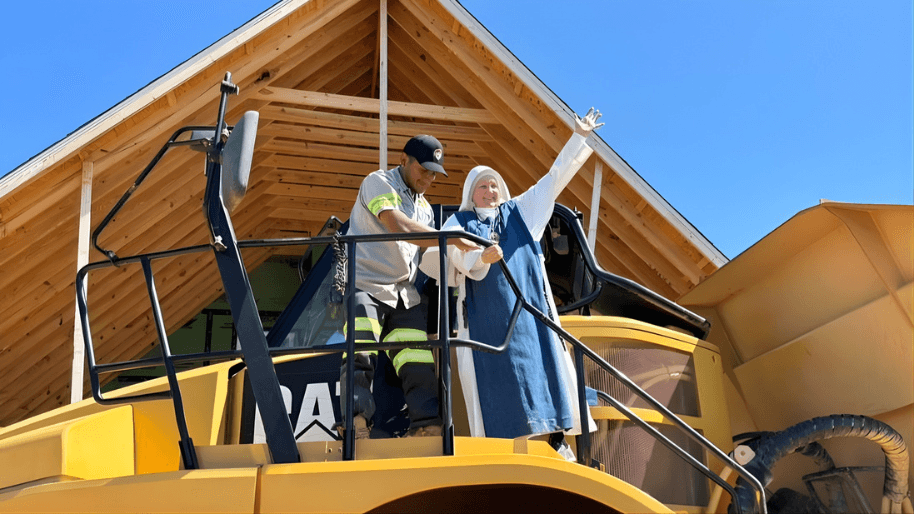 Two people standing on the steps of a large yellow excavator in front of a timber-framed building, one of them with an arm raised.