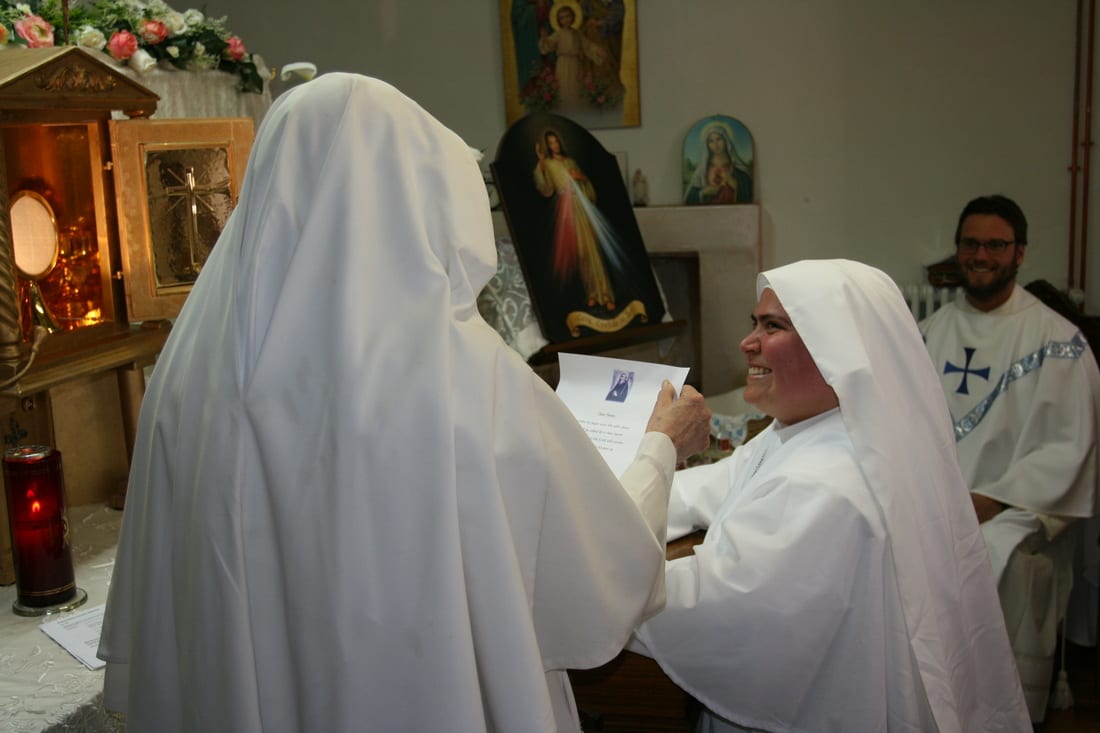 A sister in a white habit smiling as she receives a folded card from another sister, with a priest and religious icons behind them in a small chapel room.
