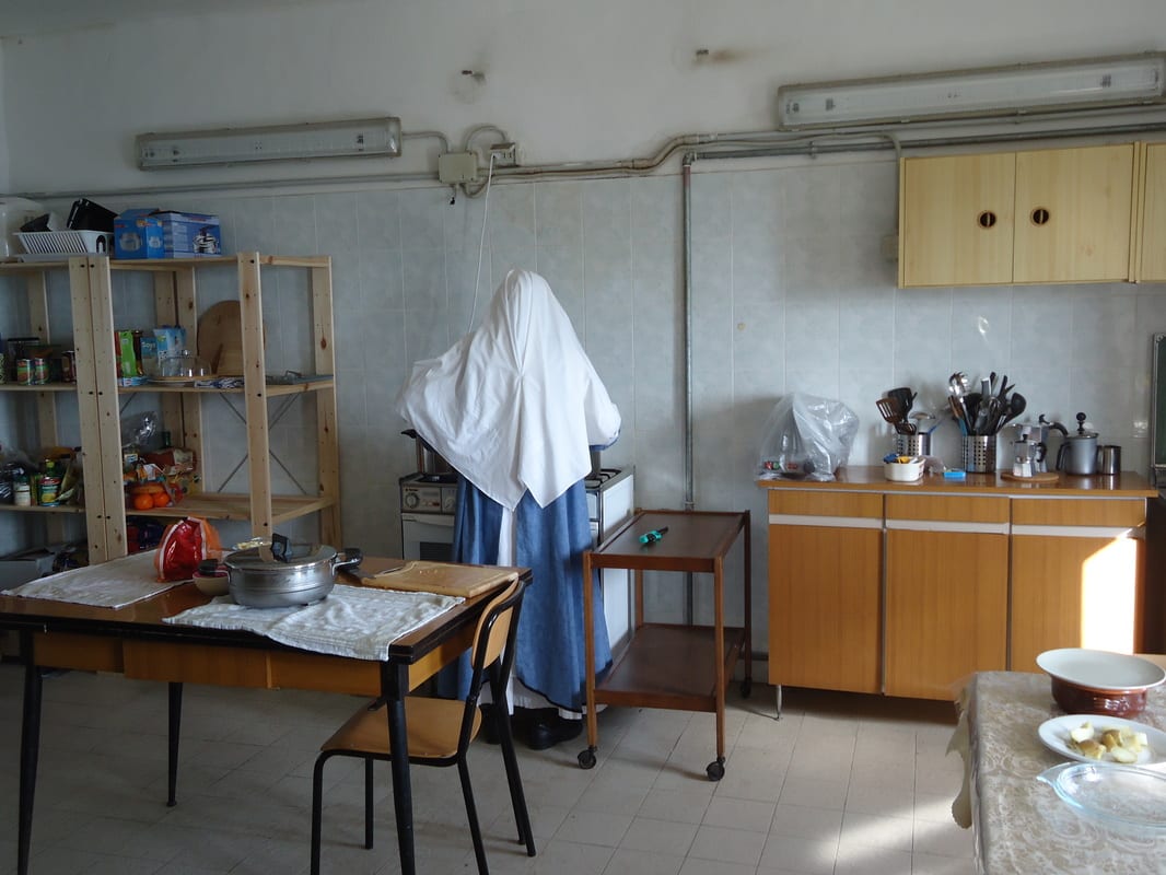 A sister in a white habit standing at a kitchen counter, back to the camera, amid basic kitchen equipment and shelving.