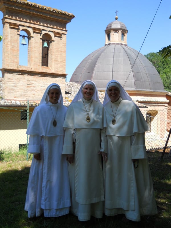 Three sisters in white habits standing outdoors in front of an Italian church with a dome and bell tower.