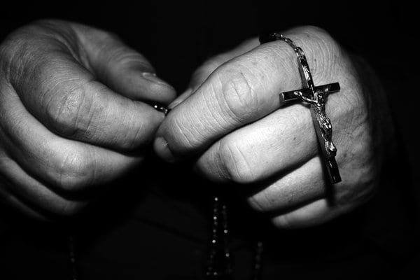 Close-up black-and-white photograph of aged hands holding a crucifix rosary in prayer.