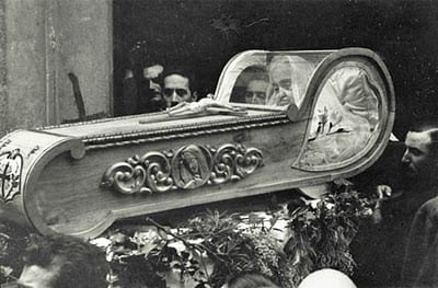 Historical photograph showing the ornate carved wooden reliquary bier of the Servant of God Luisa Piccarreta, surrounded by onlookers.