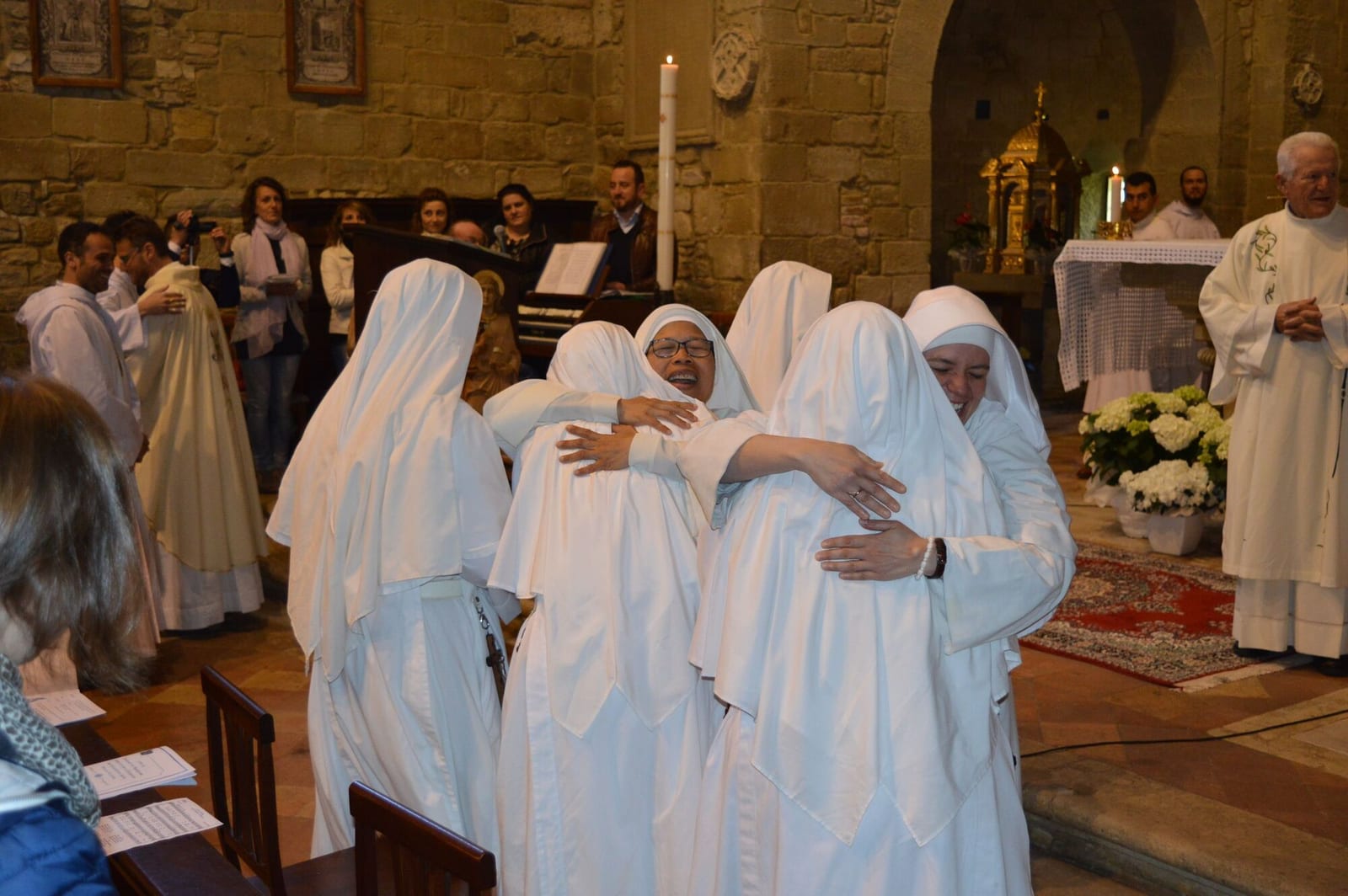 Sisters in white veils embracing one another in a stone church during a ceremony, with a priest and a congregation nearby.