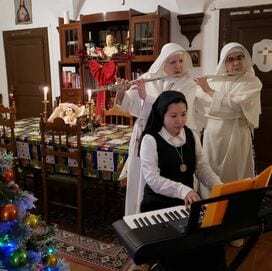 Three sisters in white habits and a young woman in a black top playing instruments together in a decorated room at Christmas time: two sisters on flute, one young woman at a keyboard.