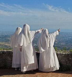 Three sisters in white habits seen from behind, standing at a stone parapet with one pointing toward a panoramic coastal landscape.