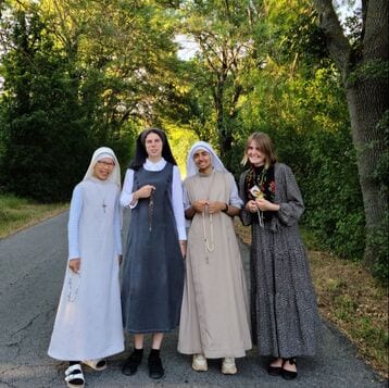 Four people standing together on a wooded road: two sisters in white habits, one in a grey habit, and a young woman in a floral dress, all holding rosaries.