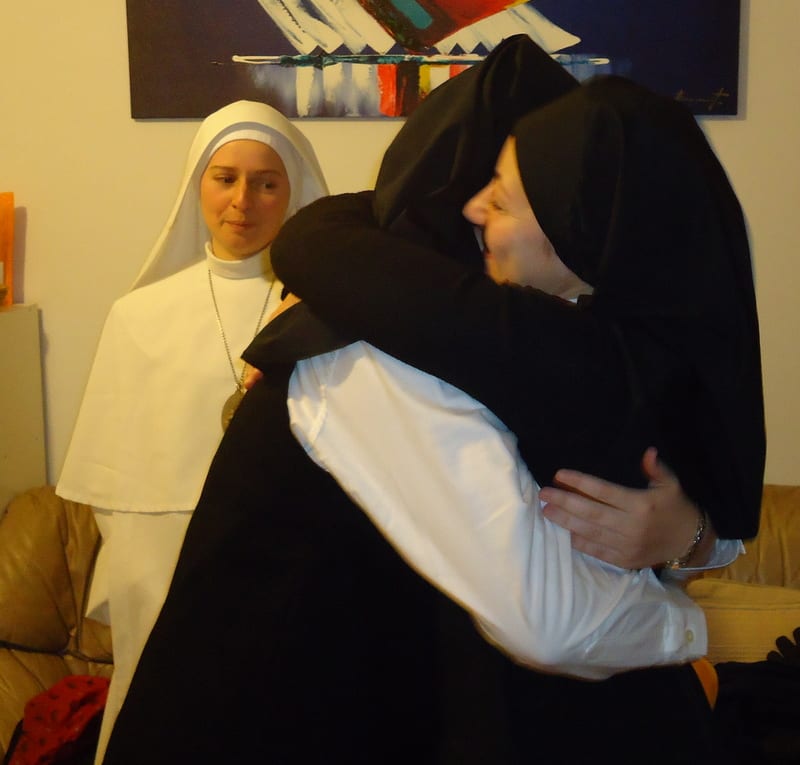 Two sisters embracing warmly; a third sister in white looks on from behind.