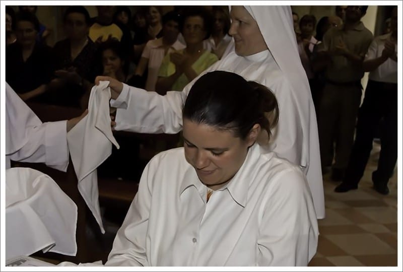 A woman bowing her head as a sister in white places a veil over her during a ceremony in a church.