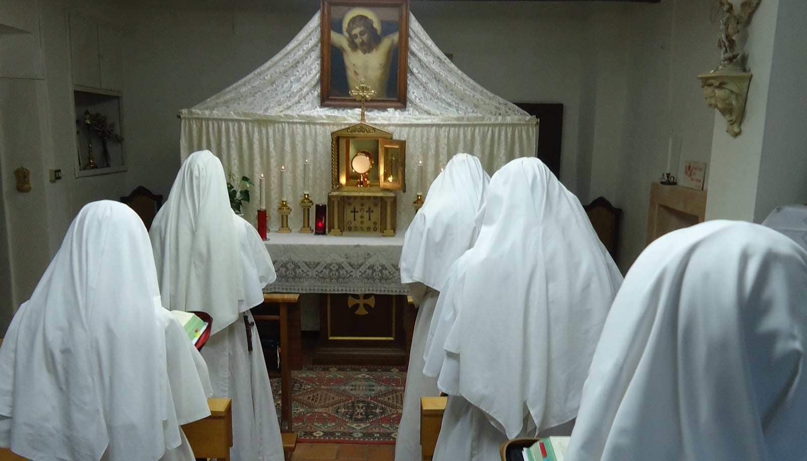 Sisters kneeling in white veils before a small altar where the Blessed Sacrament is exposed in a monstrance beneath a canopy.