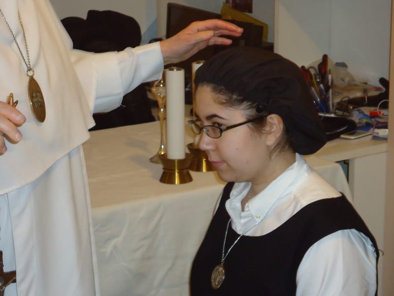 A priest placing his hand on the head of a kneeling young woman during a ceremony, with a lit candle and draped table visible.