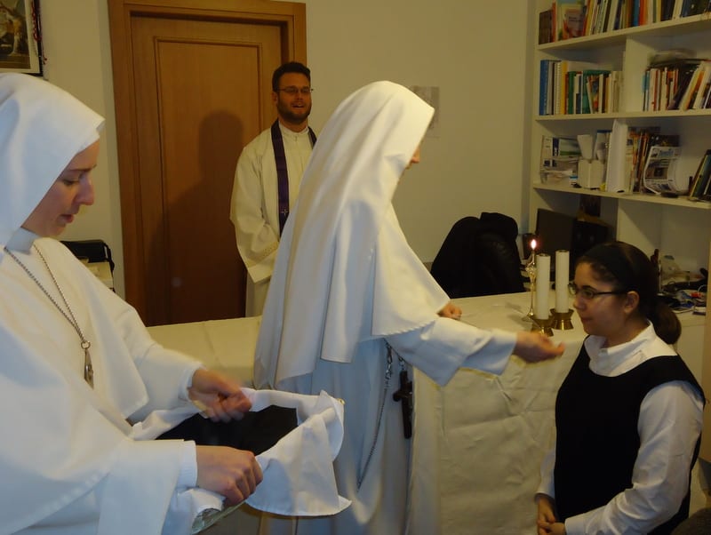 A sister presenting a book to another sister who is being received; a priest and bookshelf are visible in the background.