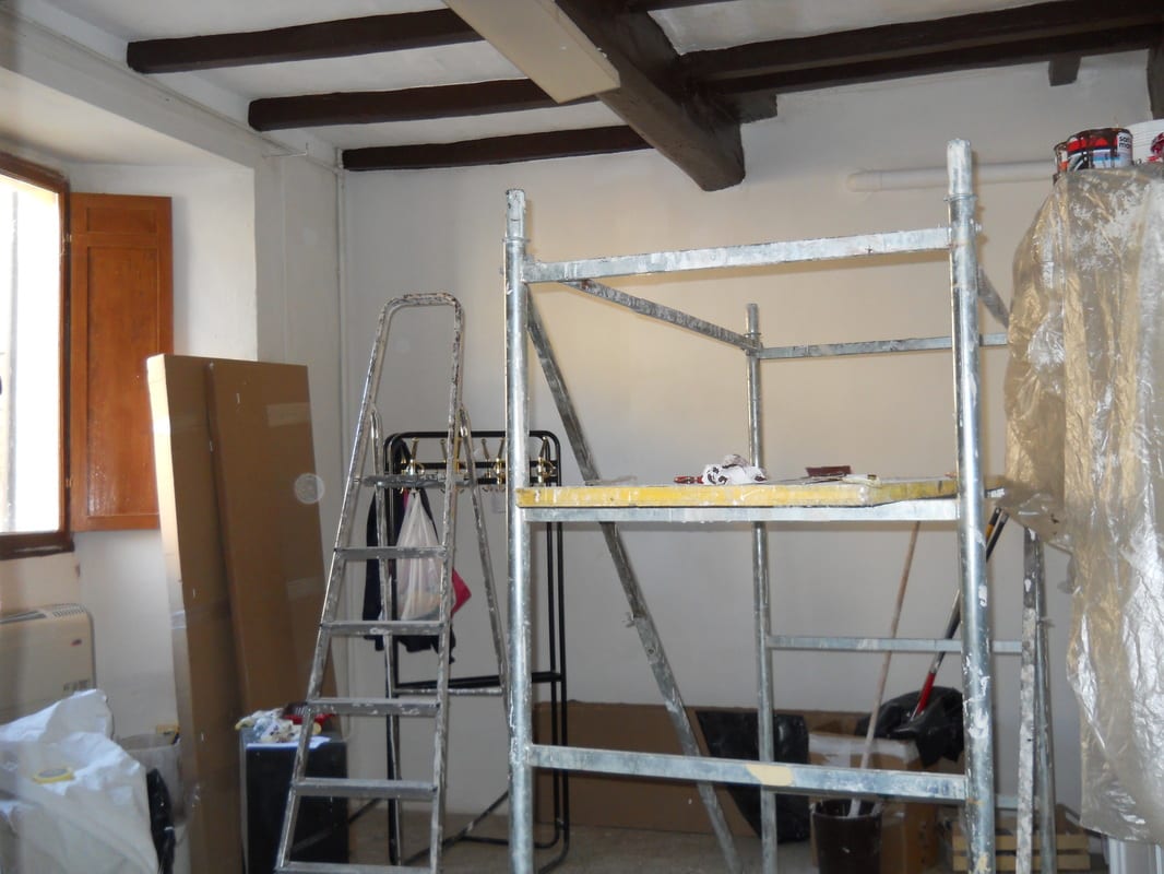 An empty room under renovation, with scaffolding erected against white walls and exposed wooden ceiling beams.