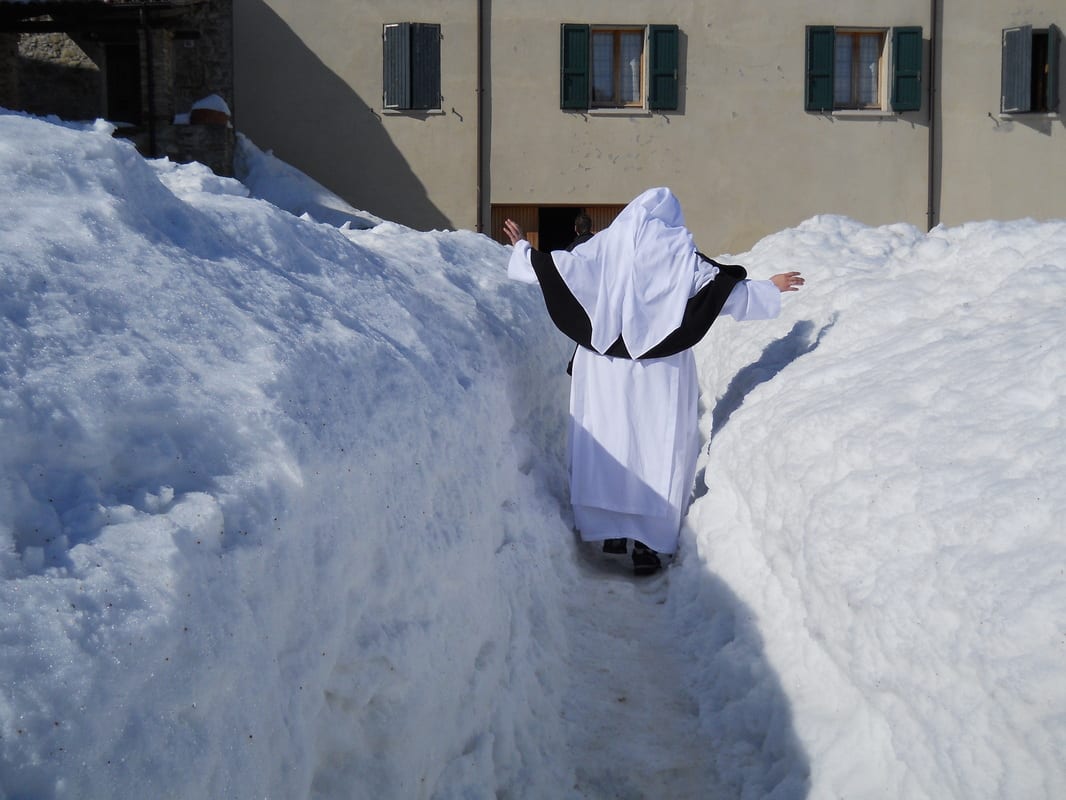 A sister in a white and black habit walking through a deep narrow path cut between walls of snow, arms outstretched.
