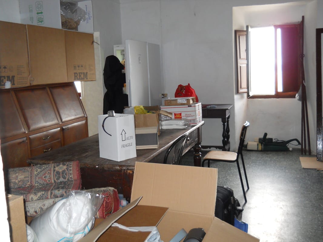 A cluttered interior room filled with cardboard boxes, loose furniture, and stacked cabinets awaiting clearing.