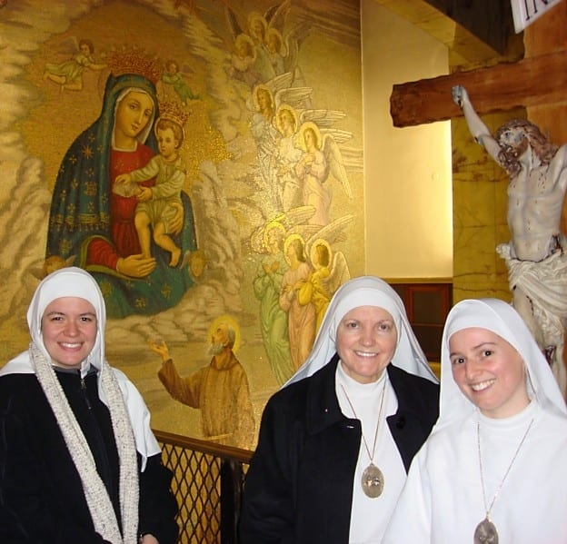 Three sisters in white habits smiling inside a chapel, with a large religious mural of Our Lady and angels on the wall behind them and a crucifix to the right.