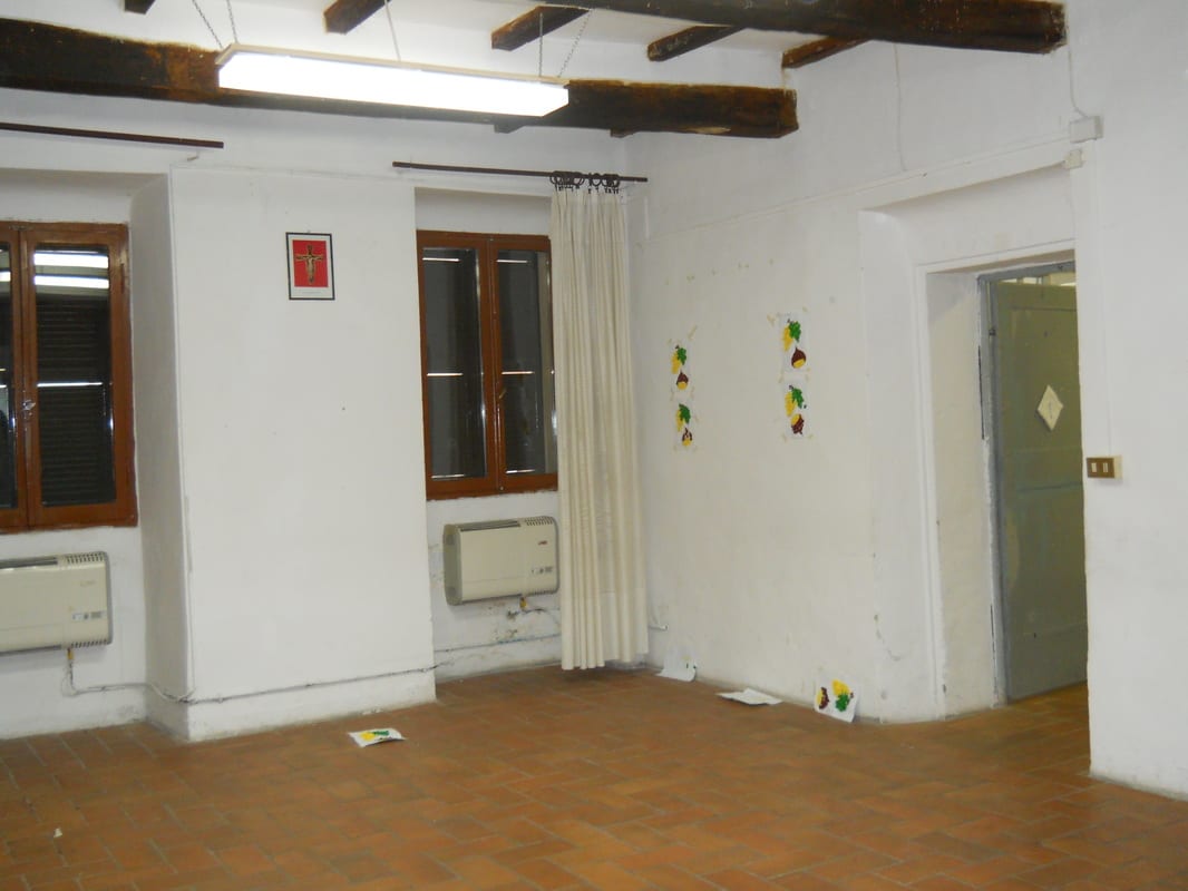 A plain empty room with fluorescent lighting, two wooden windows, bare white walls, and a door at the far end.