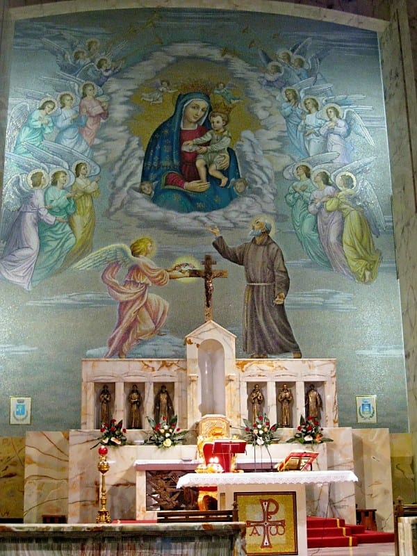 An ornate church altar with a large painted apse fresco of Our Lady with angels and a saint, flanked by fresh flower arrangements.