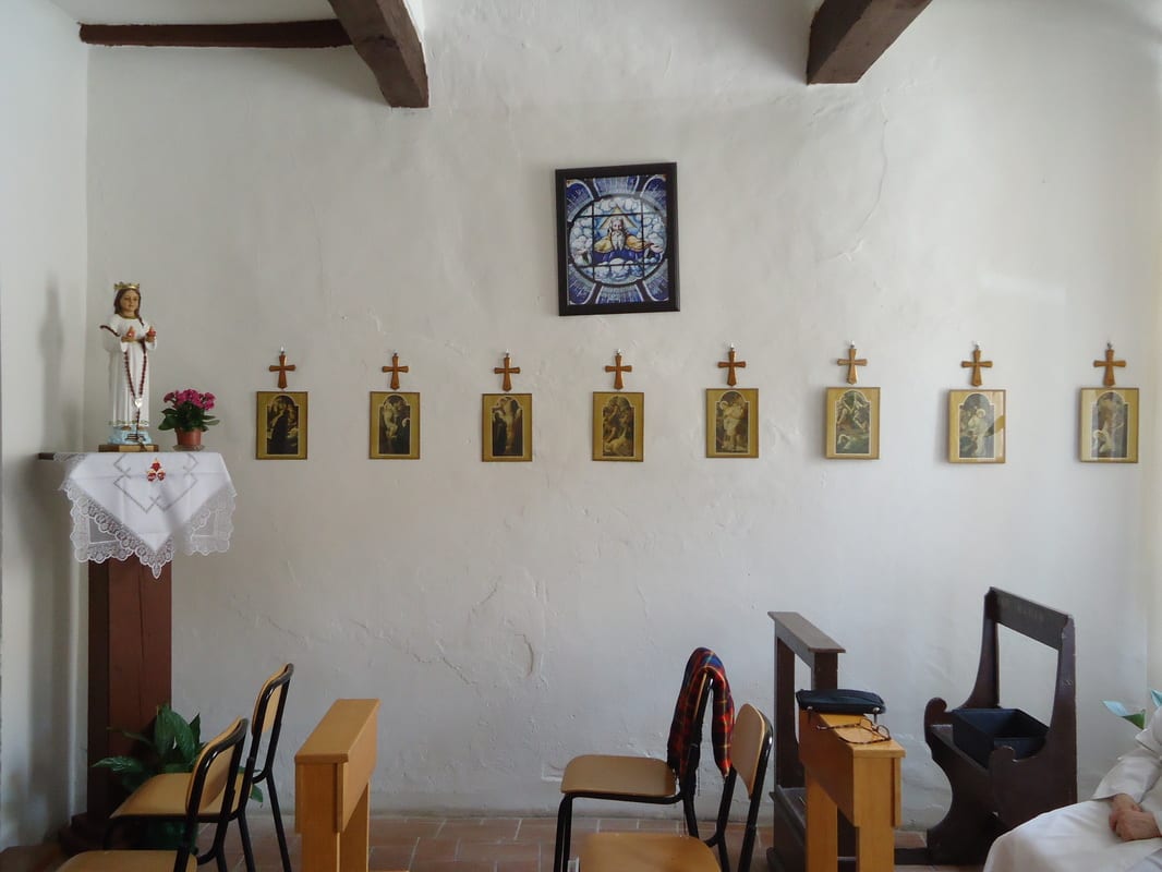 A simple whitewashed chapel interior with stations-of-the-cross icons and small crosses spaced along the wall, chairs arranged for prayer, and a small side altar.