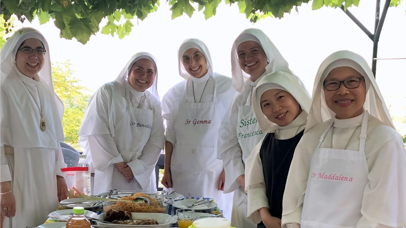 Six sisters in white habits standing behind an outdoor table of food beneath a leafy canopy.
