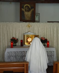 A sister in a white habit kneeling alone in prayer before a small lit monstrance on an altar, with candles and a painting of the crucified Christ above.