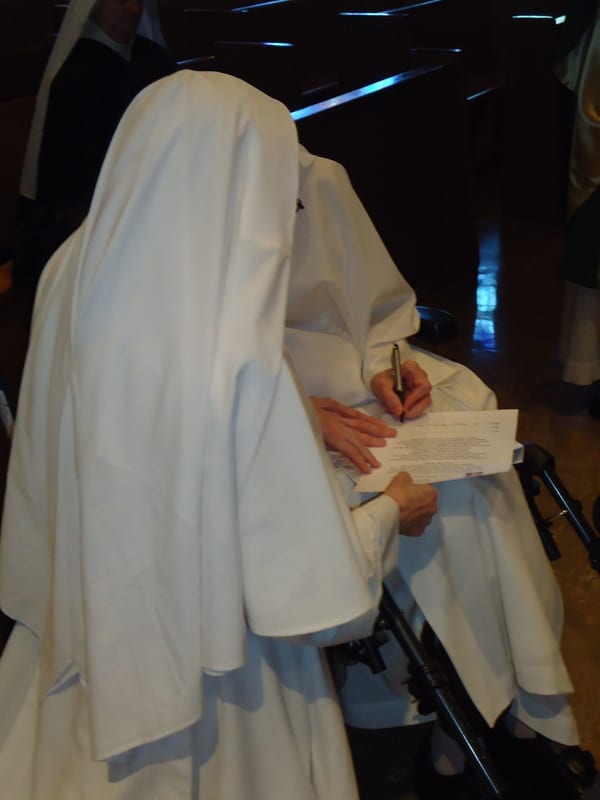A sister in a white habit bent over a pew or chair, signing a document or book.