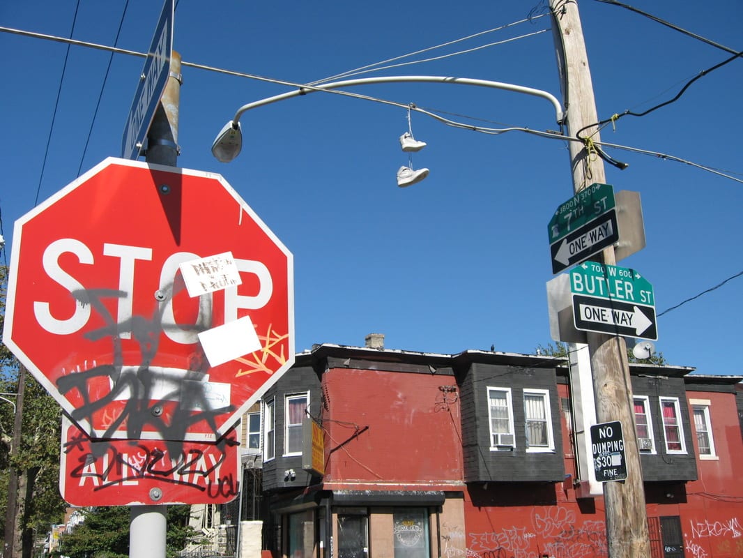 A graffiti-covered stop sign at the corner of 7th Street and Butler Street, with sneakers dangling from overhead wires and red brick row houses in the background.