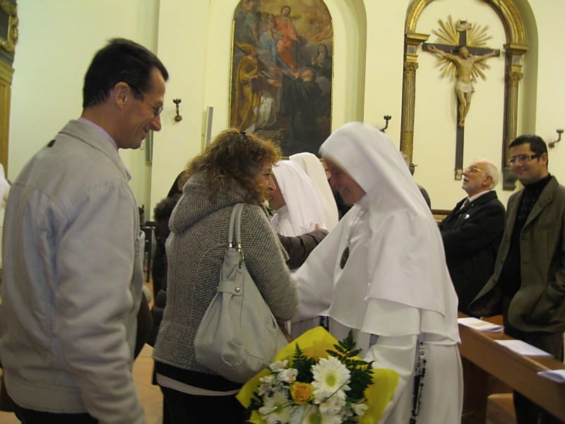 A sister in a white habit greets a woman holding flowers inside a church, with other attendees visible in the background.