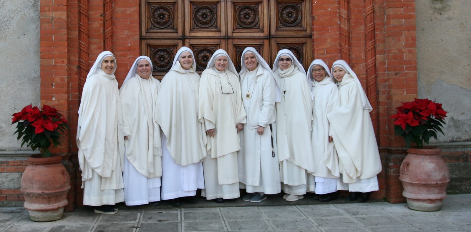 Seven sisters in white choir mantles standing before the carved wooden doors of a church, with poinsettias on either side.