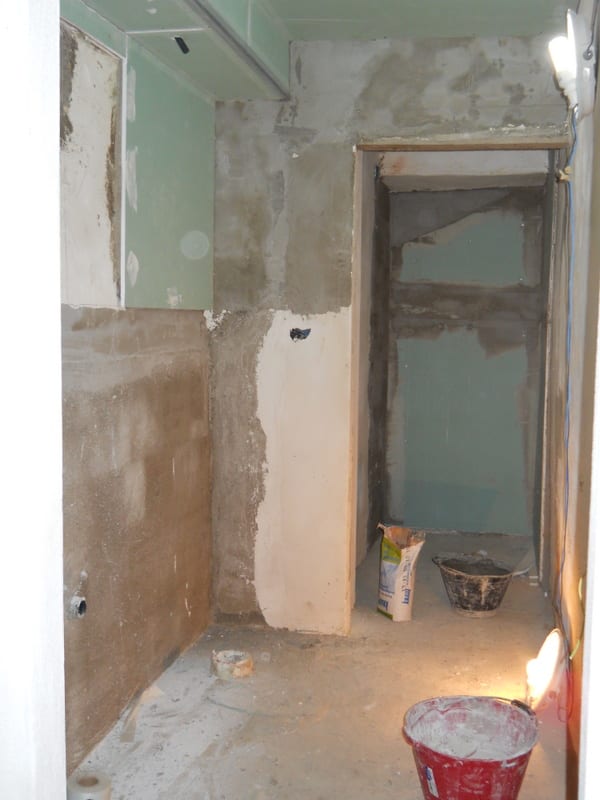 Stripped bathroom walls down to bare plaster, no fixtures, with a bucket and debris on the floor.