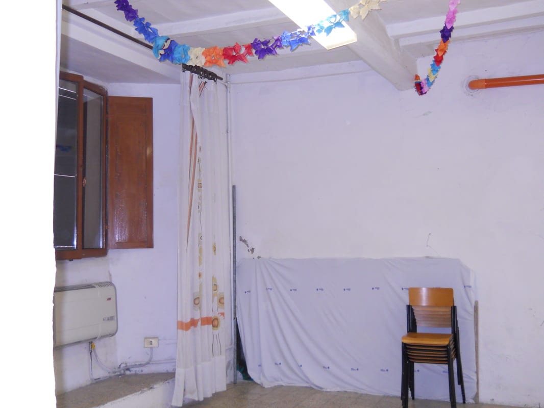 An empty room with bare white walls, paper garland strung across the ceiling, a sheer curtain, and a single wooden chair.