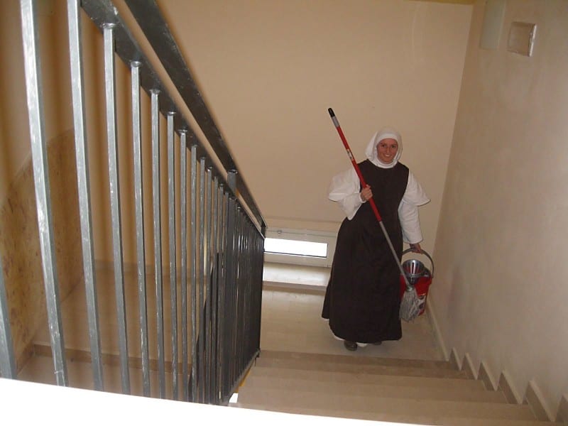 A sister in a black habit carrying a mop and bucket descends a staircase with metal banisters.
