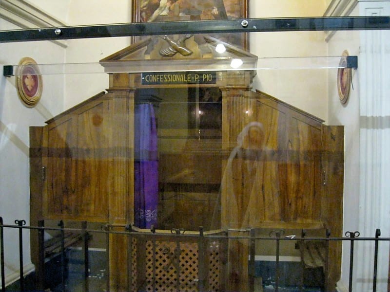A wooden confessional with the label "Confessionale-Pio" enclosed behind a glass barrier in a church.