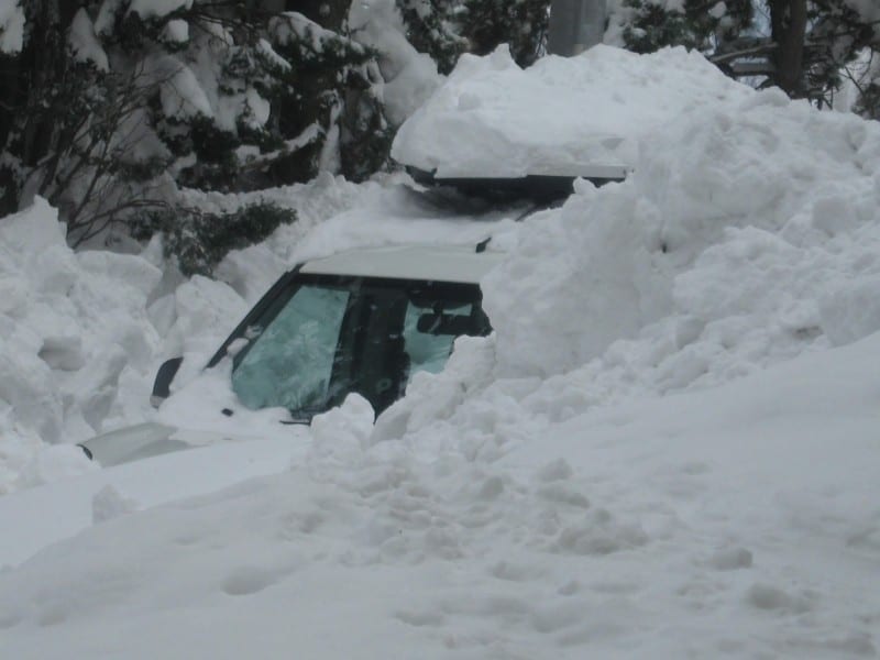 A van almost entirely buried in deep snow, its windshield and roof just visible above the drifts.