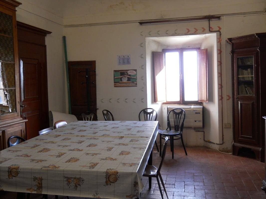 A room with a large table, mismatched chairs, a wooden sideboard, and a window with a radiator underneath.