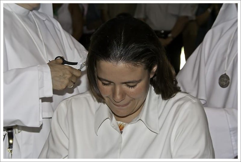 Close-up of a woman's hair being cut with scissors by hands in a white habit during the investiture rite.